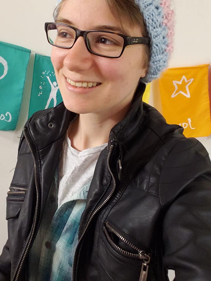 Seb smiling, dressed in a pleather jacket, button-down shirt, T-shirt and a beanie